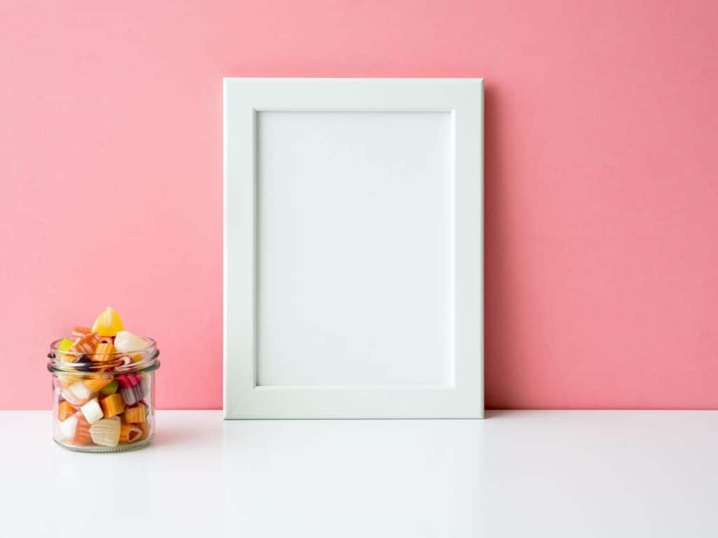 Blank white frame and lollipops in jar on a white table against the pink wall with copy space